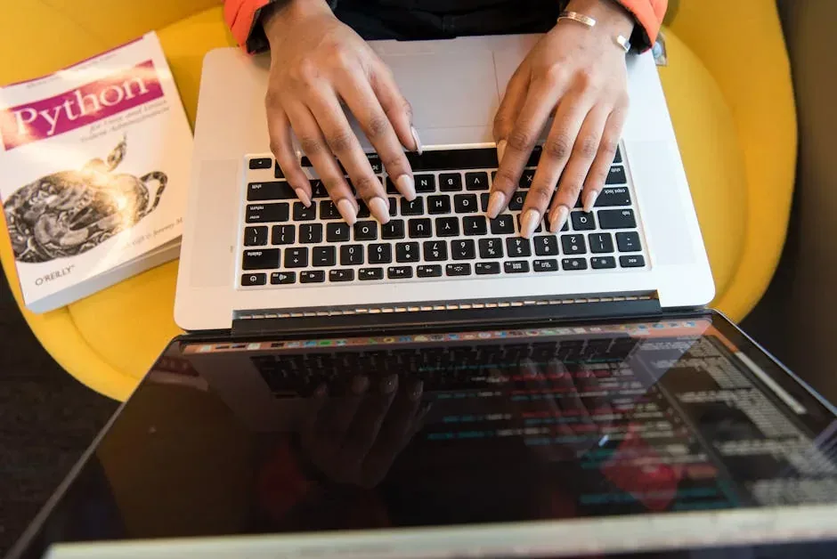 Person's hands typing on a laptop next to a Python programming book. Laptop screen displays reflected code, suggesting development.